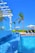 A luxurious outdoor swimming pool with cascading water and clear blue tiles, surrounded by palm trees. In the background, sun loungers with umbrellas and a gazebo can be seen on a wooden deck, creating a serene tropical atmosphere under a clear blue sky.