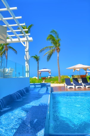 A luxurious outdoor swimming pool with cascading water and clear blue tiles, surrounded by palm trees. In the background, sun loungers with umbrellas and a gazebo can be seen on a wooden deck, creating a serene tropical atmosphere under a clear blue sky.