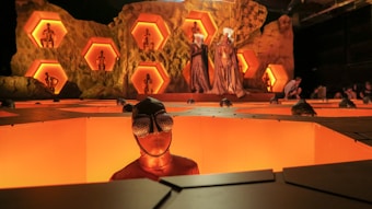 A futuristic scene featuring individuals wearing textured costumes, with one person prominently in the foreground wearing bug-eyed glasses. The backdrop includes a rocky structure with hexagonal alcoves, each containing figures in poses. The lighting creates a warm, orange glow, contributing to the surreal atmosphere.