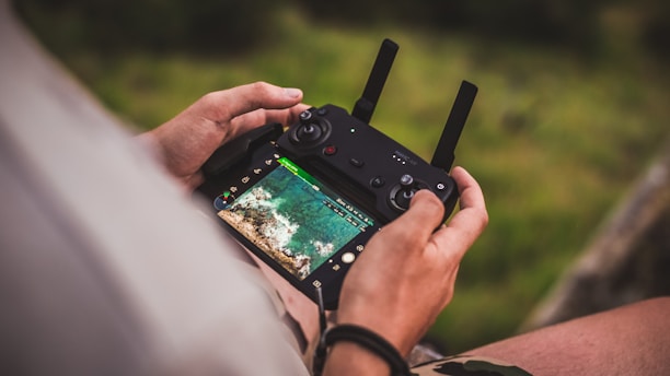 A person is holding a drone remote control with a screen displaying an aerial view of a landscape. The image focuses closely on the hands and the controller, with the lush green background softly blurred.