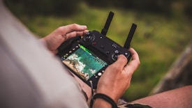 A person is holding a drone remote control with a screen displaying an aerial view of a landscape. The image focuses closely on the hands and the controller, with the lush green background softly blurred.