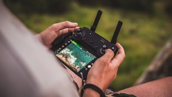 A person is holding a drone remote control with a screen displaying an aerial view of a landscape. The image focuses closely on the hands and the controller, with the lush green background softly blurred.