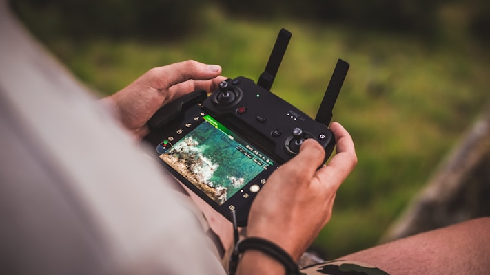 A person is holding a drone remote control with a screen displaying an aerial view of a landscape. The image focuses closely on the hands and the controller, with the lush green background softly blurred.