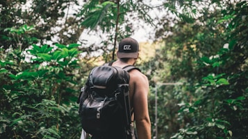 A person with a black backpack is walking through a lush jungle path surrounded by dense green foliage. The individual is wearing a black cap and tank top, and their back is turned towards the viewer.