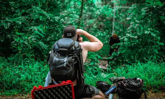 A person with a backpack is seated in a lush green forest, looking through binoculars. They are surrounded by vibrant foliage, with another person in the background appearing to be on a swing or zip line. Several bags and equipment are scattered on the ground.