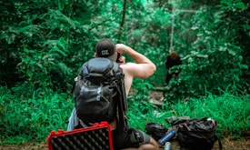 A person with a backpack is seated in a lush green forest, looking through binoculars. They are surrounded by vibrant foliage, with another person in the background appearing to be on a swing or zip line. Several bags and equipment are scattered on the ground.