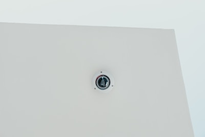 A security camera is mounted on a flat white ceiling, centrally positioned, with a minimalist and clean appearance.