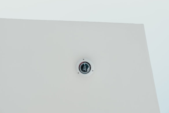 A security camera is mounted on a flat white ceiling, centrally positioned, with a minimalist and clean appearance.