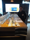 A person stands beside a conference or event registration table. The table is filled with water bottles, name tags with colored clips, pens, and papers. Behind the person, a large screen displays digital content. The room is dimly lit with some ceiling lights.