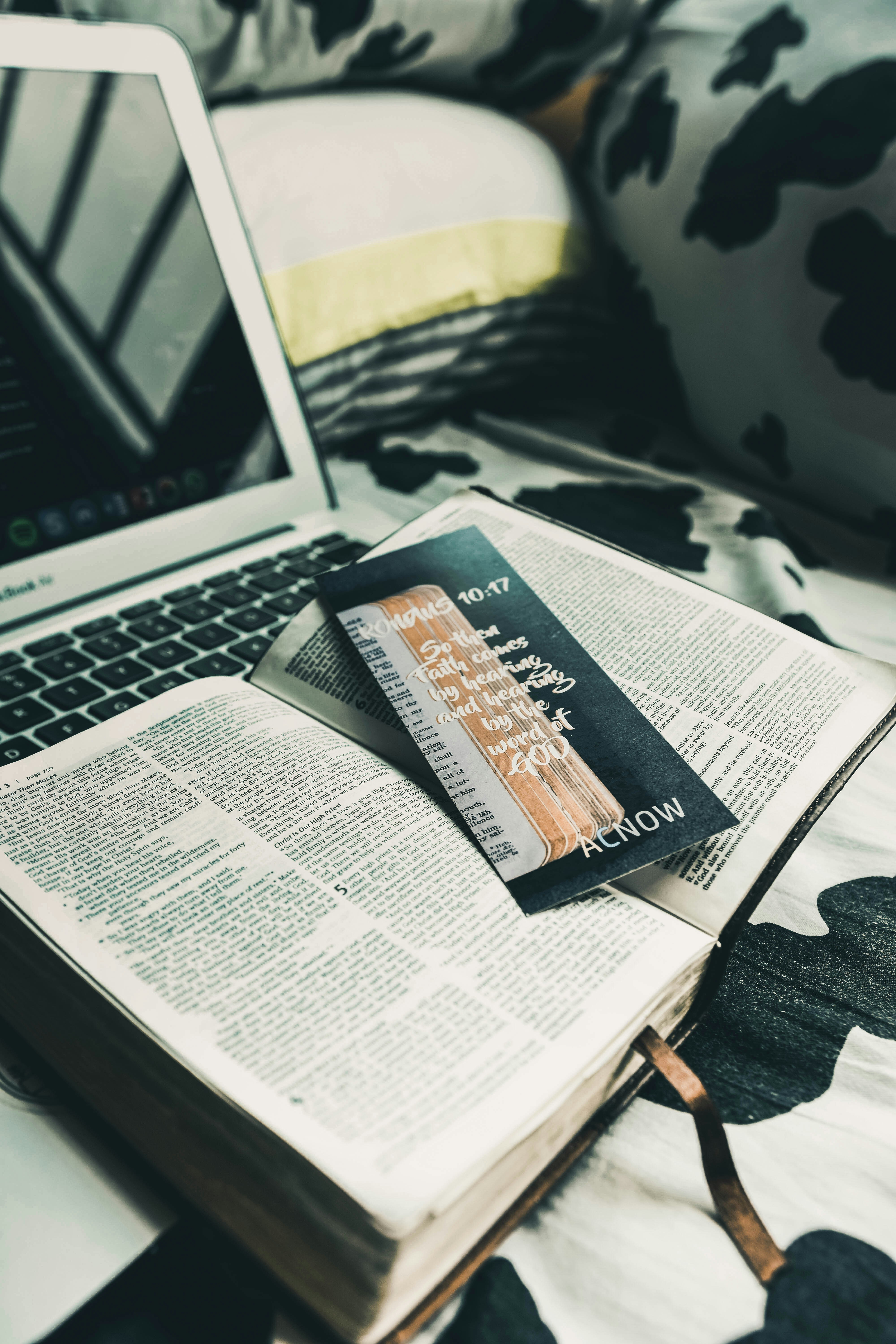 An open book is placed on a bed with black and white patterned sheets. A laptop is nearby, partially visible, with its keyboard and screen. A black bookmark with handwritten text and a design is lying across the book's pages. The setting suggests a cozy and relaxed atmosphere.