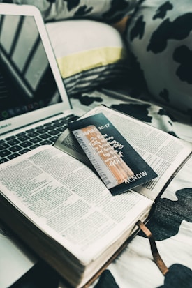 An open book is placed on a bed with black and white patterned sheets. A laptop is nearby, partially visible, with its keyboard and screen. A black bookmark with handwritten text and a design is lying across the book's pages. The setting suggests a cozy and relaxed atmosphere.