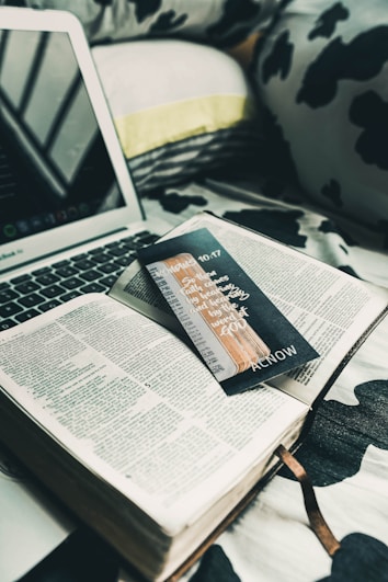 An open book is placed on a bed with black and white patterned sheets. A laptop is nearby, partially visible, with its keyboard and screen. A black bookmark with handwritten text and a design is lying across the book's pages. The setting suggests a cozy and relaxed atmosphere.