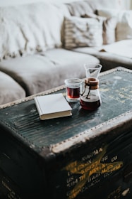 A rustic wooden table or chest with a stained surface holds a glass container of red liquid, a clear glass, and a closed notebook. In the background, a soft, light gray couch with textured pillows adds a cozy ambiance.