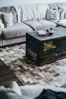 A cozy living room setup featuring a gray sectional sofa with cushions and a cream-colored throw blanket. In front of the sofa, a rustic black trunk serves as a coffee table. On top of the trunk, there are two glasses of red liquid and a closed white book. The floor is adorned with a textured, geometric-patterned rug.