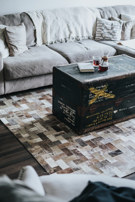 A cozy living room setup featuring a gray sectional sofa with cushions and a cream-colored throw blanket. In front of the sofa, a rustic black trunk serves as a coffee table. On top of the trunk, there are two glasses of red liquid and a closed white book. The floor is adorned with a textured, geometric-patterned rug.