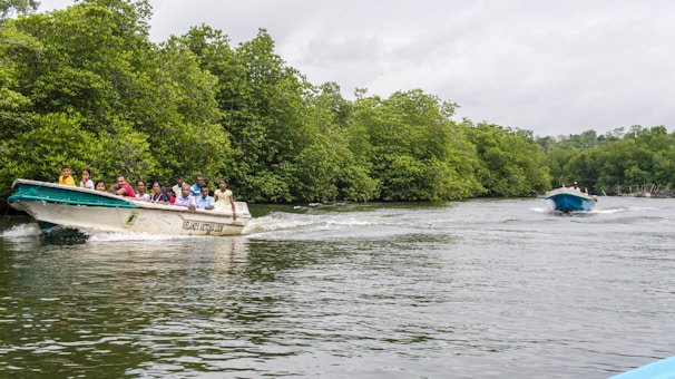 A group of people are traveling in a motorboat along a river surrounded by lush green trees. Another boat is seen in the distance on the right side of the river. The water is calm and reflects the overcast sky above.