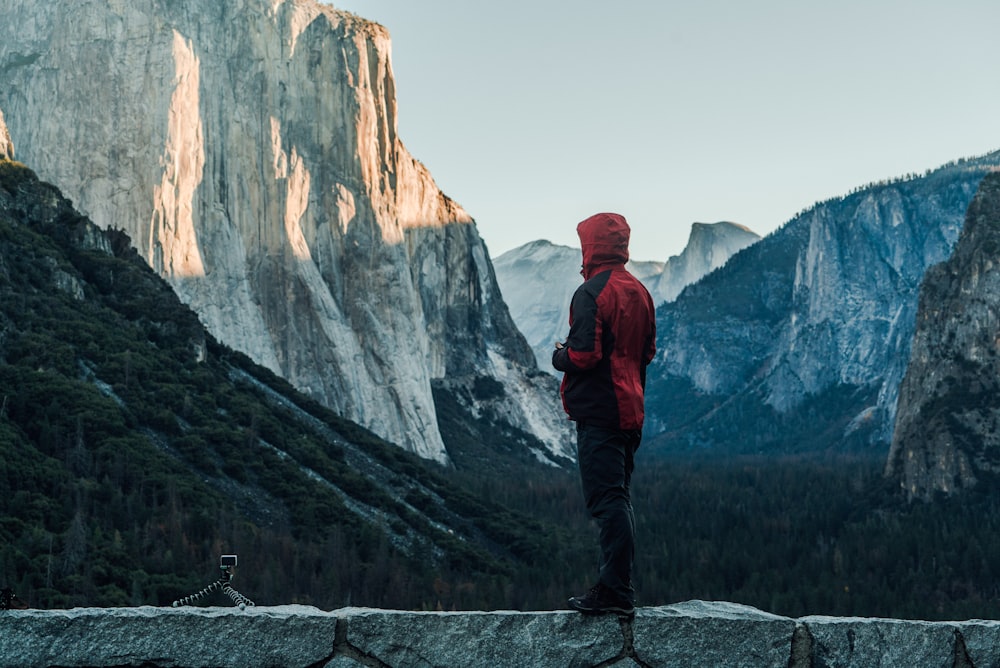 person wearing red hooded jacket standing on rock facing of yosemite park