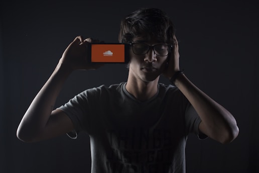 A person with glasses is holding a smartphone next to their head, with the screen displaying an orange icon. The lighting creates a dramatic, low-key atmosphere, emphasizing shadows and contrasts against a dark backdrop.