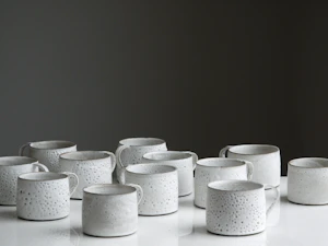 Rows of white speckled ceramic mugs on a white surface