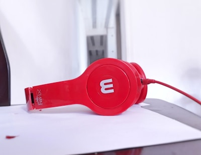 A pair of red over-ear headphones lies on a white surface. The headphones feature a prominent round ear cup with the letter 'M' displayed prominently in white. A matching red cable is connected to the headphones, and the environment appears minimalistic with bright lighting.