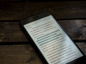 A smartphone placed on a wooden table, displaying a passage of text that appears to be religious in nature. The focus is on the screen highlighting the text in a dimly lit environment, creating a serene and contemplative atmosphere.