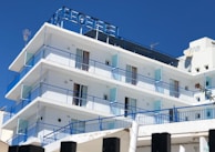 A multi-story hotel building with a clear blue sky backdrop. The structure features several balconies with blue railings and glass panels. The exterior is predominantly white, and the top displays an antenna and satellite dish. The word 'HOTEL' is prominently displayed at the rooftop section.