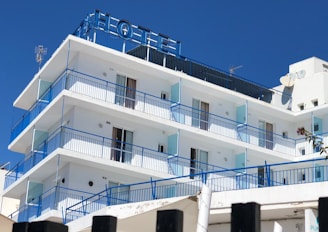 A multi-story hotel building with a clear blue sky backdrop. The structure features several balconies with blue railings and glass panels. The exterior is predominantly white, and the top displays an antenna and satellite dish. The word 'HOTEL' is prominently displayed at the rooftop section.
