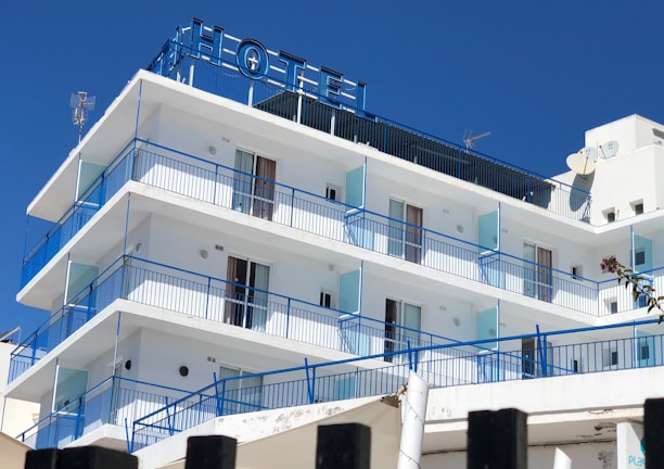 A multi-story hotel building with a clear blue sky backdrop. The structure features several balconies with blue railings and glass panels. The exterior is predominantly white, and the top displays an antenna and satellite dish. The word 'HOTEL' is prominently displayed at the rooftop section.