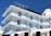 A multi-story hotel building with a clear blue sky backdrop. The structure features several balconies with blue railings and glass panels. The exterior is predominantly white, and the top displays an antenna and satellite dish. The word 'HOTEL' is prominently displayed at the rooftop section.