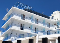 A multi-story hotel building with a clear blue sky backdrop. The structure features several balconies with blue railings and glass panels. The exterior is predominantly white, and the top displays an antenna and satellite dish. The word 'HOTEL' is prominently displayed at the rooftop section.