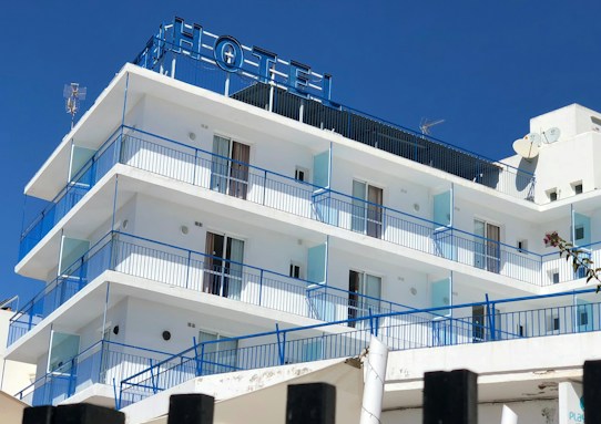 A multi-story hotel building with a clear blue sky backdrop. The structure features several balconies with blue railings and glass panels. The exterior is predominantly white, and the top displays an antenna and satellite dish. The word 'HOTEL' is prominently displayed at the rooftop section.