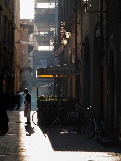 A narrow street flanked by old buildings with a vintage aesthetic. A person in a white apron stands on the left side, surrounded by soft light and shadows. Bicycles are parked near a small ristorante with plants on top. Street lamps and neon signs hang from the walls, while scaffolding is visible at the end of the street.