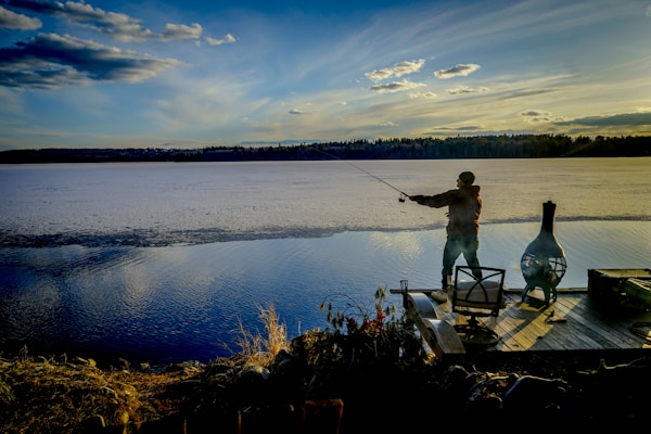 A person is fishing off a wooden dock with a rod, overlooking a calm, expansive lake under a wide, blue sky filled with scattered clouds. The distant shoreline is lined with trees, and the scene is bathed in the warm, golden light of twilight. Near the person, there is a chiminea and some outdoor furniture.
