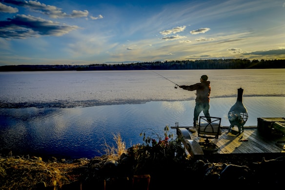 A person is fishing off a wooden dock with a rod, overlooking a calm, expansive lake under a wide, blue sky filled with scattered clouds. The distant shoreline is lined with trees, and the scene is bathed in the warm, golden light of twilight. Near the person, there is a chiminea and some outdoor furniture.