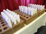 Several rolled-up white papers are neatly arranged in wooden holders with circular slots. Each roll appears to be tied with a light blue ribbon. The holders sit on a white tablecloth, and a maroon curtain serves as the background.