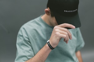 A person wearing a black cap with the words 'Grace Chapel' emblazoned on it. They are tilting their head down slightly, with a hand adjusting or holding the brim of the cap. The individual is also wearing a light green shirt and a black wristband with white text on it. The background is a muted gray tone, providing a neutral setting that emphasizes the subject.