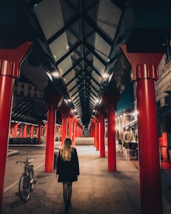 A person with long blonde hair walks under a structure with bright red columns and a geometric glass roof. The setting appears urban, possibly a city street or walkway, with a bicycle parked on the side. The scene is illuminated with artificial lights, creating shadows and highlighting the architectural design.