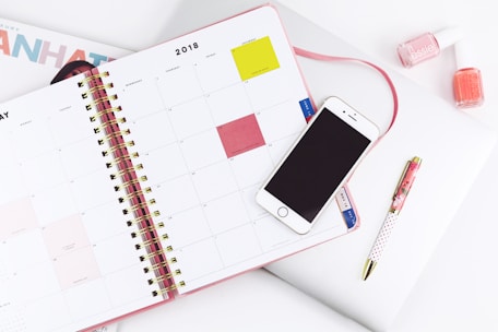 An open planner displaying a monthly calendar with a smartphone placed on top. Nearby are a pen and two bottles of nail polish. The setup suggests a workspace or study area with attention to organization and style.