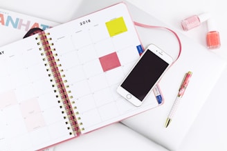 An open planner displaying a monthly calendar with a smartphone placed on top. Nearby are a pen and two bottles of nail polish. The setup suggests a workspace or study area with attention to organization and style.
