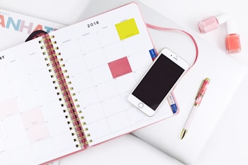 An open planner displaying a monthly calendar with a smartphone placed on top. Nearby are a pen and two bottles of nail polish. The setup suggests a workspace or study area with attention to organization and style.