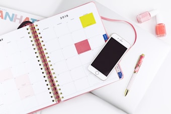An open planner displaying a monthly calendar with a smartphone placed on top. Nearby are a pen and two bottles of nail polish. The setup suggests a workspace or study area with attention to organization and style.