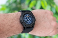 A close-up of an arm wearing a smart watch with a black strap, displaying various health and activity metrics including a step counter and heart rate monitor, set against a blurred green leaf background.