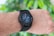 A close-up of an arm wearing a smart watch with a black strap, displaying various health and activity metrics including a step counter and heart rate monitor, set against a blurred green leaf background.