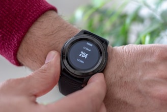 A close-up view of a person's wrist wearing a black smartwatch with a touchscreen displaying fitness statistics such as steps, calories burned, and distance. The person is wearing a red sweater and is interacting with the watch using their other hand.