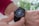 A close-up view of a person's wrist wearing a black smartwatch with a touchscreen displaying fitness statistics such as steps, calories burned, and distance. The person is wearing a red sweater and is interacting with the watch using their other hand.