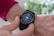 A close-up view of a person's wrist wearing a black smartwatch with a touchscreen displaying fitness statistics such as steps, calories burned, and distance. The person is wearing a red sweater and is interacting with the watch using their other hand.
