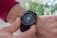 A close-up view of a person's wrist wearing a black smartwatch with a touchscreen displaying fitness statistics such as steps, calories burned, and distance. The person is wearing a red sweater and is interacting with the watch using their other hand.