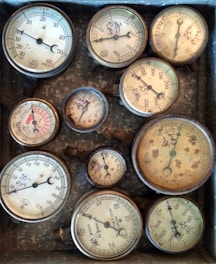 Several vintage pressure gauges with different dials and scales are arranged within a worn metallic box. The gauges vary in size, showing wear and age, with some markings and numbers handwritten on the glass surface.