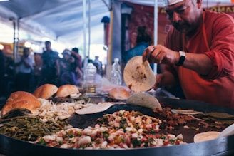 A man is cooking at a street food market, preparing a variety of ingredients on a large hot plate, including tortillas, grilled vegetables, and meats. The scene is bustling with activity in the background, indicating a lively atmosphere.