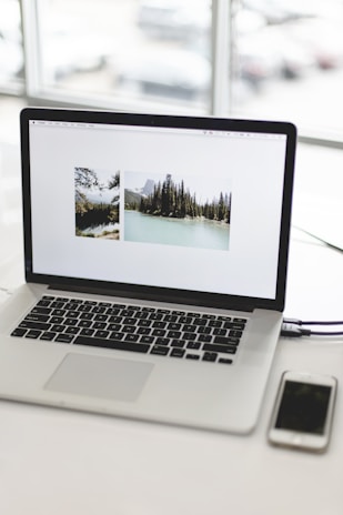 A laptop on a desk displaying two nature photographs on its screen. The wooden desk contrasts with the modern tech. An iPhone is placed next to the laptop. The background features large windows with blurry views of cars.